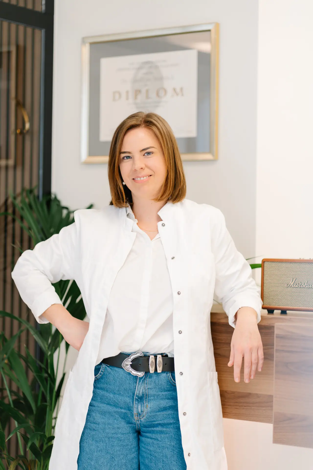 OÄ Dr. Caroline Stieböck, Wahlarzt und Augenarzt Bad Goisern, lächelt in einem weißen Kittel und Jeans. Im Hintergrund das Diplom.